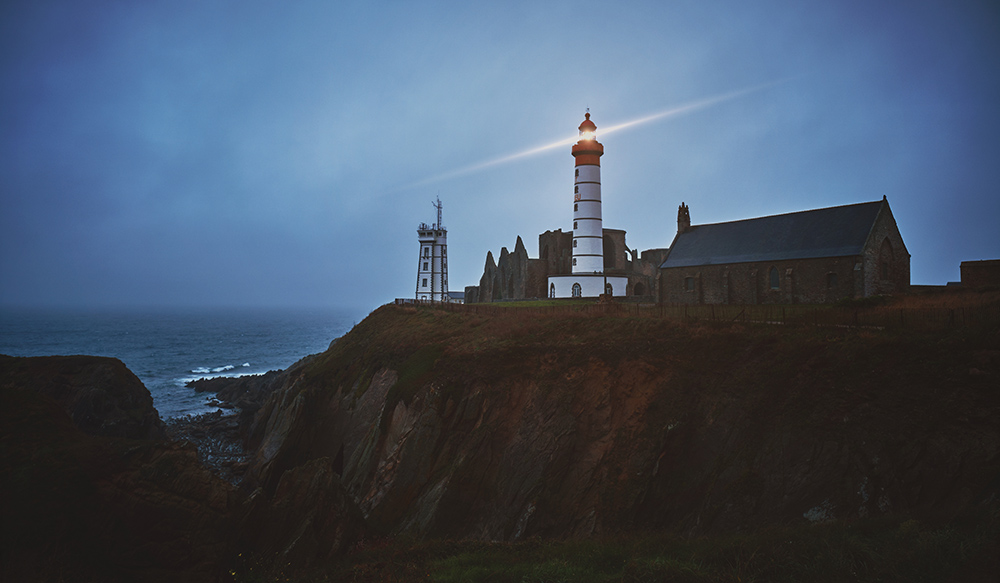 Horizontal shot of a mysterious town on a cliff with a white turned-on lighthouse during dusk